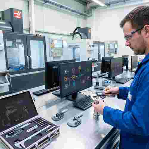 Techniker prüft Werkstück mit Messschieber in moderner CNC-Werkstatt; CNC-Maschinen im Hintergrund.