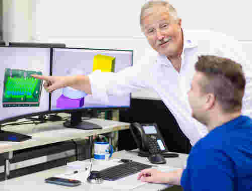 Ein älterer Mann mit grauem Haar und Schnauzbart zeitg auf einen PC-Bildschirm. Neben ihm sitzt ein junger Mann. Auf dem Bildschirm sidn Abläufer der CAD/CAM Technologie zu sehen.