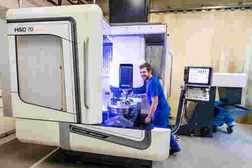 Ein junger Mann in blauer Kleidung an einer hochmodernen CNC-Fräsmaschine von Werkzeugtechnik Schmitz. Diese steht an einer Wand, im Hintergrund ist ein PC zu erkennen.