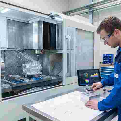 Moderne CNC-Fräsmaschine fräst Metallteil, Späne fliegen; Techniker prüft Messschieber, Tablet