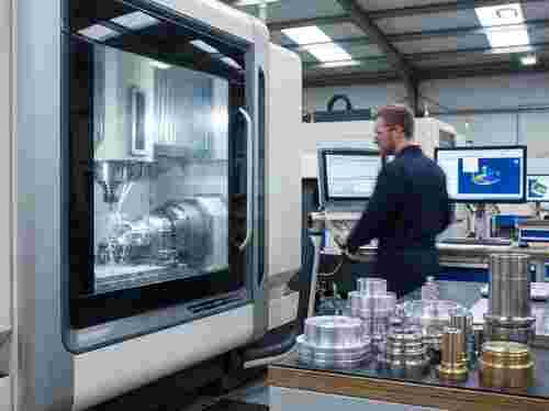 Moderne CNC-Fräswerkstatt: 3–5 Achsen, Metallteile, Kühlmittel, Späne, Techniker im Hintergrund.