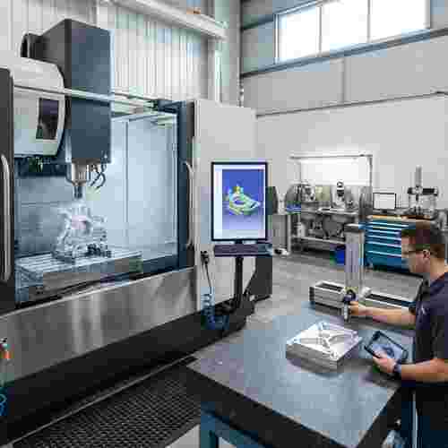 Quadratisches Bild einer modernen Fräswerkstatt mit 5-Achs-CNC, Bauteil geprüft, CAD/CAM-Monitor.