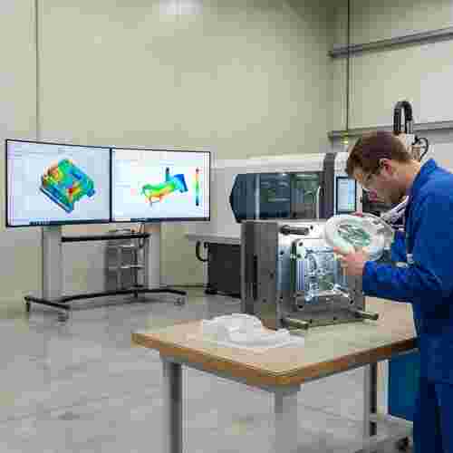 Techniker prüft Metallwerkzeug in moderner Spritzgusswerkstatt vor CAD- und Simulationsbildschirmen.